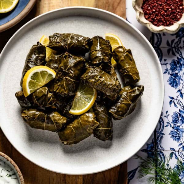 A plate piled with Turkish stuffed grape leaves garnished with lemon slices, served with a bowl of yogurt sauce.