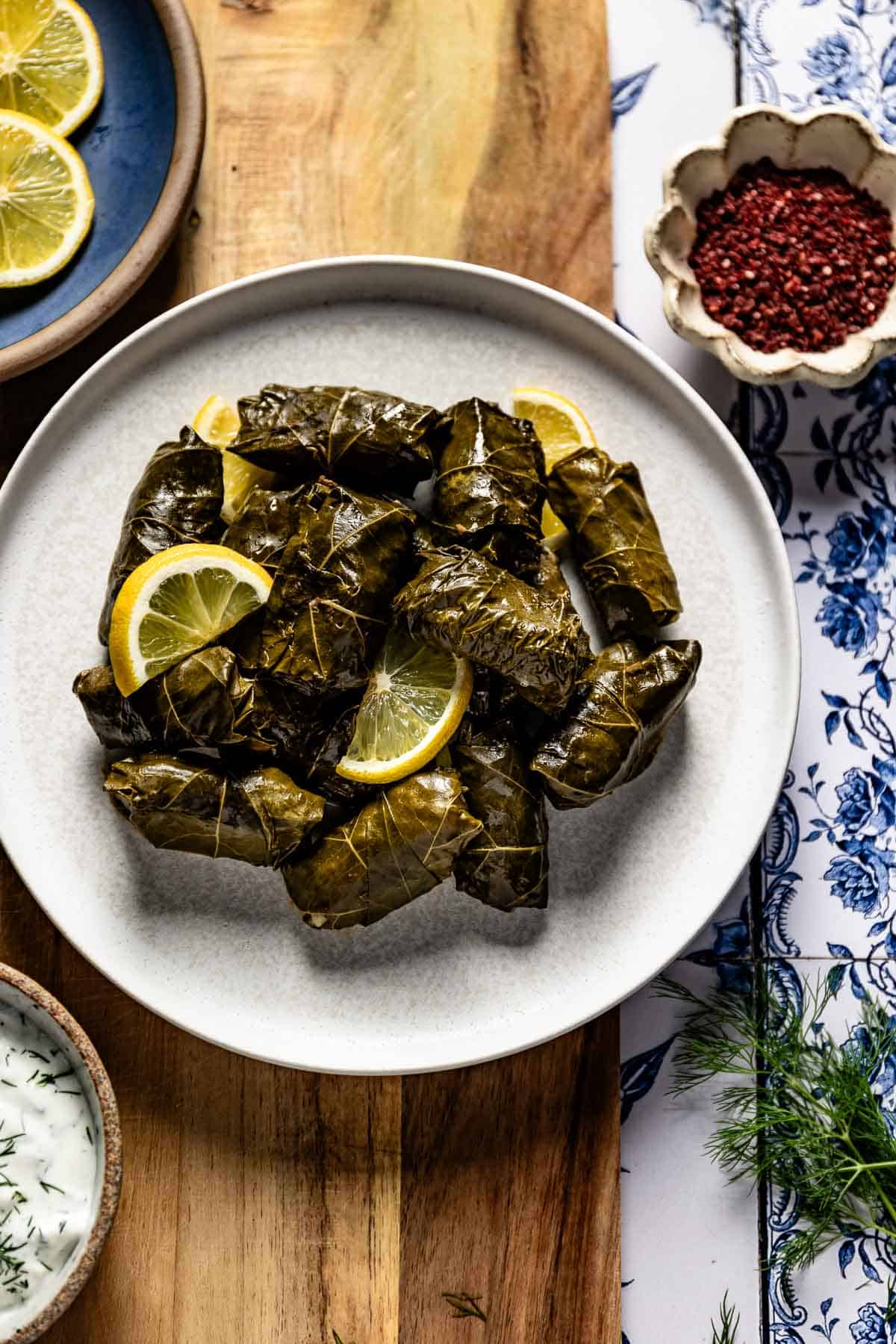 A plate piled with Turkish stuffed grape leaves garnished with lemon slices, served with a bowl of yogurt sauce.
