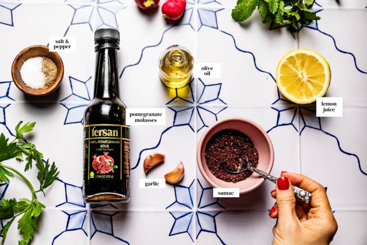 Fattoush dressing ingredients on a blue tiled surface: pomegranate molasses, olive oil, lemon, garlic, salt and pepper, and a pink bowl of sumac.