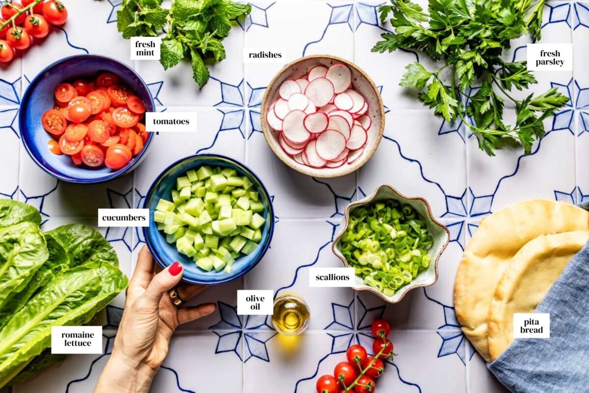 Fattoush salad ingredients laid out on blue tile: romaine, cherry tomatoes, cucumbers, radishes, scallions, fresh mint and parsley, and pita bread.
