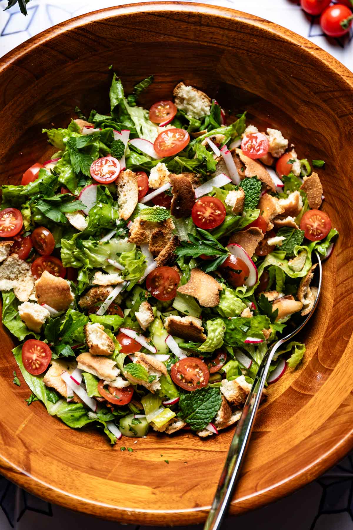 Fattoush salad in a wooden bowl with crisp pita, cherry tomatoes, radishes, romaine, and mint, with a serving spoon.