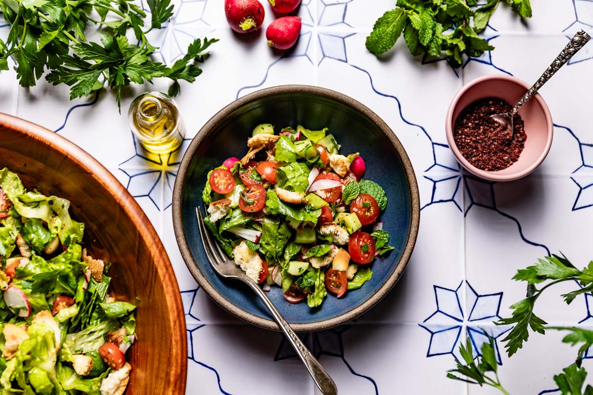 A single serving of fattoush salad in a blue bowl with a fork, surrounded by parsley, radishes, mint, olive oil, and sumac.