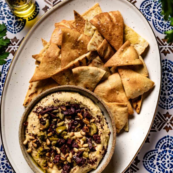 Golden homemade pita chips piled beside a bowl of hummus topped with olives and pine nuts.