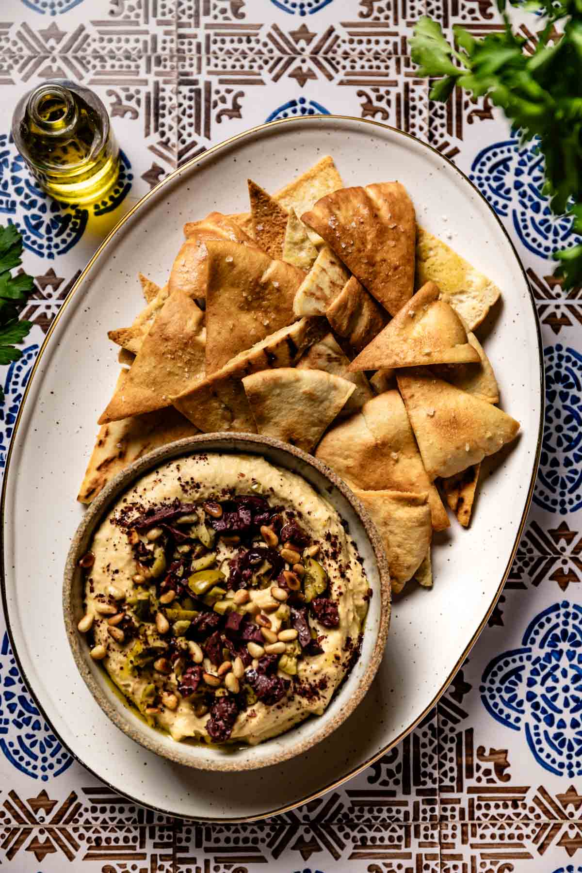 Golden homemade pita chips piled beside a bowl of hummus topped with olives and pine nuts.