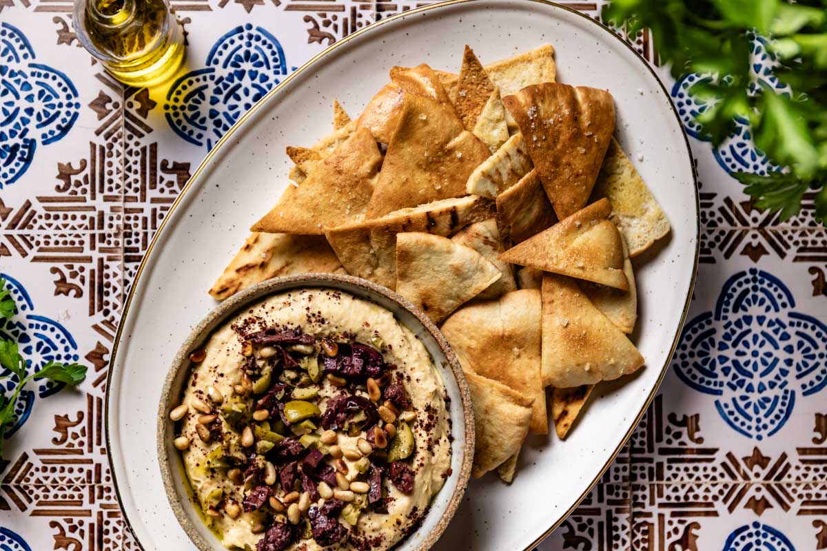 Baked pita chips arranged around a bowl of Mediterranean hummus on a patterned tile surface.
