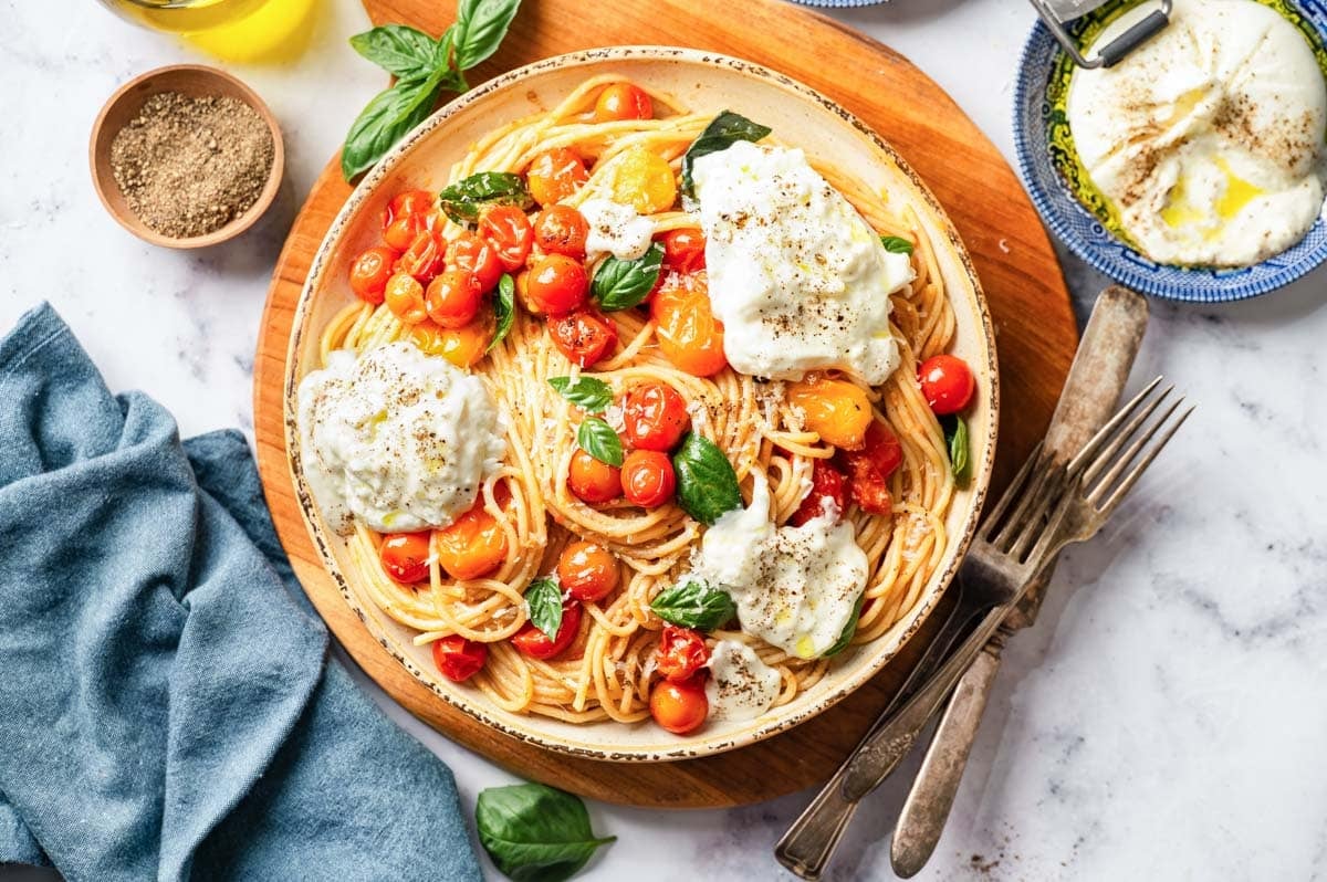 A bowl of creamy burrata pasta with burst cherry tomatoes on a wooden board from the top view.