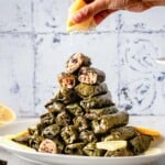 A person is squeezing lemon juice over stuffed grape leaves piled on a plate like a tower.