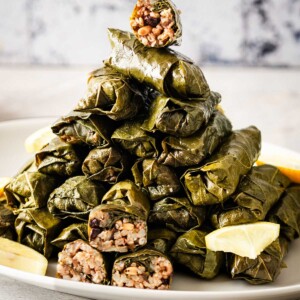 Vegetarian stuffed grape leaves piled on a plate from the front view with wedges of lemon on the side.