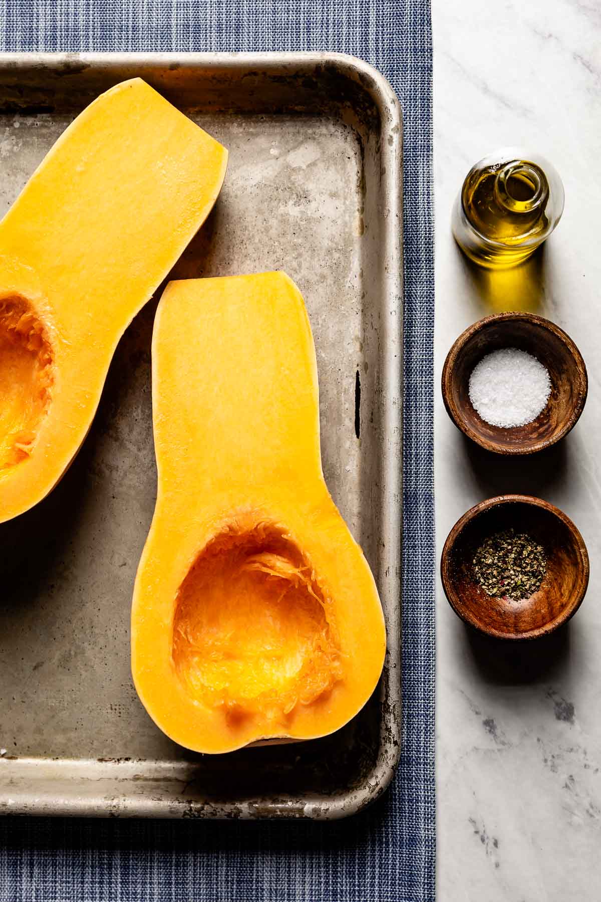 Two raw butternut squash halves, seeds scooped out, on a sheet pan next to olive oil, kosher salt, and black pepper.