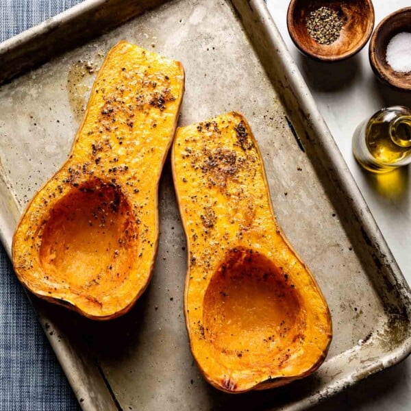 Roasted butternut squash halves, cut side up, seasoned with salt and pepper on a sheet pan, with olive oil and spices nearby.