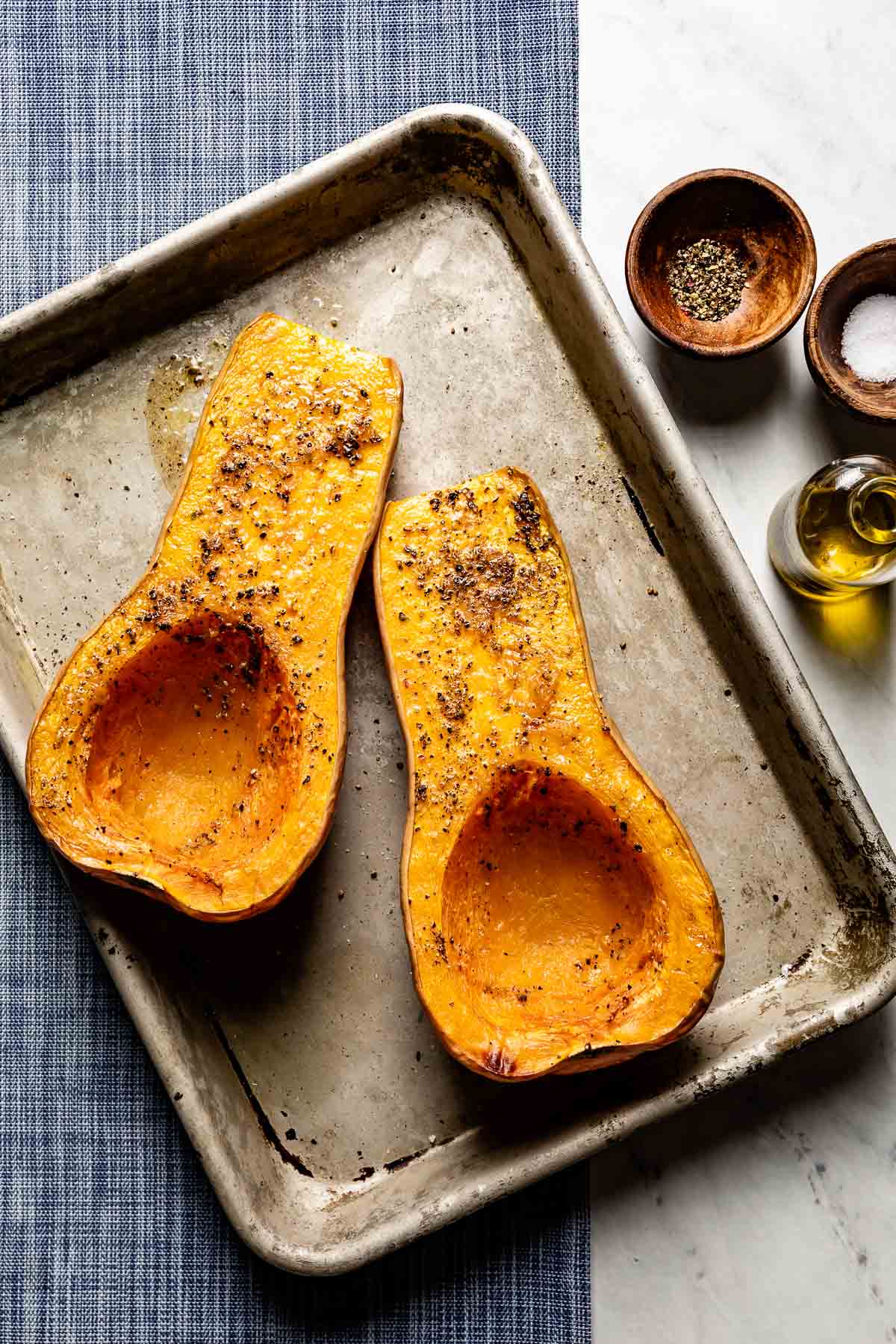 Roasted butternut squash halves, cut side up, seasoned with salt and pepper on a sheet pan, with olive oil and spices nearby.